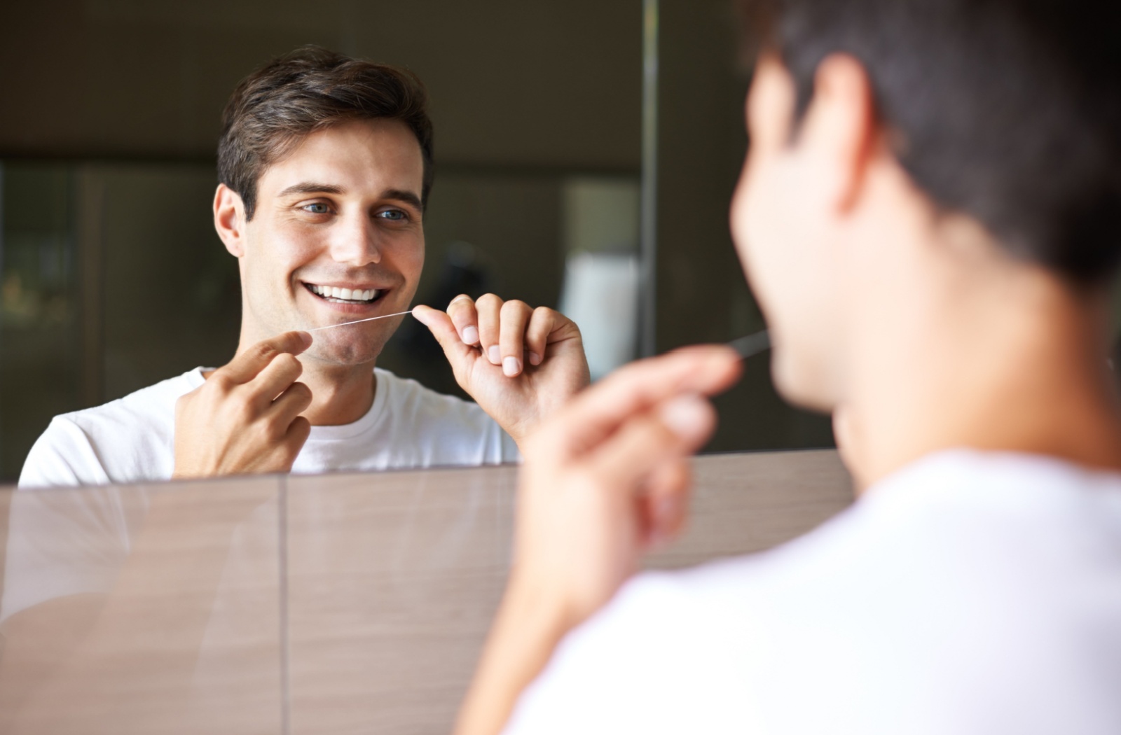 A person looking into a mirror and smiling while holding a piece of dental floss between both hands.